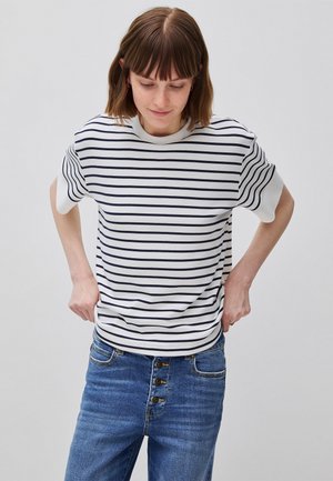 Jong persoon met schouderlang bruin haar, gekleed in een wit T-shirt met korte mouwen en zwarte strepen, en een blauwe spijkerbroek, die de tailleband van de broek aanpast.