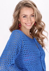 Kék horgolt felső, laza, nyitott szövésű kialakítással, kerek nyakkal és megkötős záródással. A modellen élénk rózsaszín kiegészítők és nagy fülbevalók találhatók.