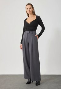 Γυναίκα ποζάροντας με μαύρη μπλούζα τύπου wrap, γκρι παντελόνι σε φαρδιά γραμμή, μαύρες μπότες έως τον αστράγαλο και χρυσά δαχτυλίδια αυτιών, μπροστά σε απλό φόντο.