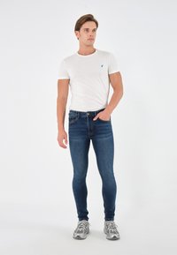 Camiseta blanca de cuello redondo con un pequeño logotipo azul, combinada con unos pantalones vaqueros ajustados azul oscuro y zapatillas deportivas grises sobre un fondo liso.