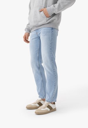 Osoba ubrana w jasnoniebieskie dżinsy, beżowo-białe trampki oraz szarą bluzę z kapturem, stojąca na białym tle z jedną ręką w kieszeni.