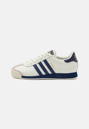 Fehér sneaker sötétkék részletekkel és három csíkkal, velúr orrésszel és texturált bőrből készült felső résszel. "SAMOA" arany betűkkel.