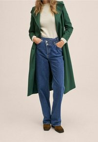 Grøn lang frakke, lysegrøn top, højtaljede blå jeans og brune sko. Frakken har en bred krave og sidelommer.