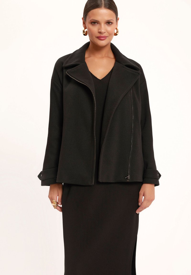 Czarny, strukturalny płaszcz z szerokim kołnierzem, asymetrycznym zapięciem na zamek oraz skośnym przodem. Teksturowany materiał z akcentami na mankietach.