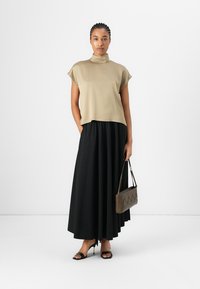 Top beige a collo alto con maniche corte, abbinato a una gonna maxi plissettata nera. La modella tiene in mano una borsa marrone con texture e indossa sandali neri.