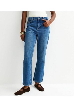 Femme portant un jean bleu taille haute à jambes droites, un haut blanc, des bracelets dorés et des ballerines Mary Jane marron, debout devant un fond blanc.