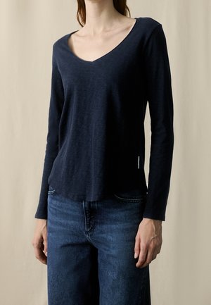 Femme portant un top uni bleu marine à manches longues avec col en V, rentré dans un jean bleu taille mi-haute sur un fond neutre.