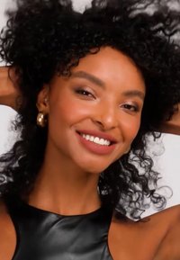 Ung kvinna med svart lockigt hår ler, bär guldörhängen och en svart ärmlös topp, händerna vilande bakom huvudet.