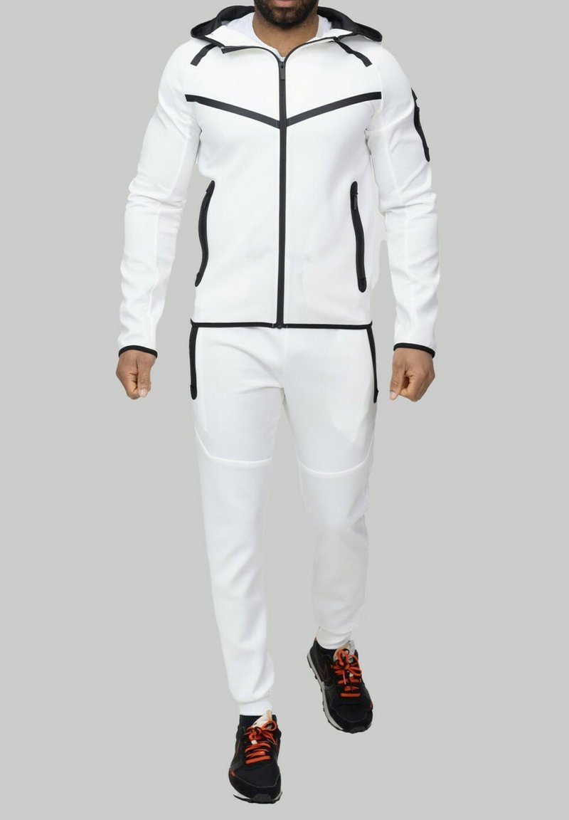 Survêtement de sport blanc avec des accents et des détails noirs, comprenant une veste à capuche et un pantalon fuselé, associé à des chaussures de course noires.