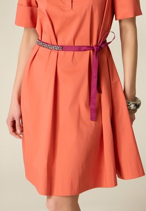 Mujer que lleva un vestido coral hasta la rodilla con mangas cortas, un cinturón fino morado con detalle de piedra preciosa y dos pulseras gruesas en la muñeca derecha.