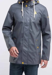 Grå huvjacka i regnmaterial med vita dragsko och gula knappar, bärs över blåjeans.