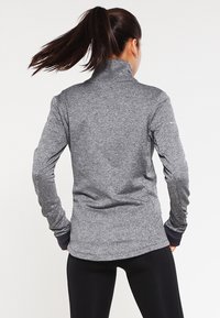 Kobieta z ciemnym kucykiem, ubrana w szary, długi rękaw sportowy top i czarne legginsy, stoi z rękami na biodrach, przodem do tyłu.