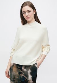 Kremowy sweter z wysokim kołnierzem, prążkowaną fakturą i opuszczonymi ramionami, zestawiony z ciemnymi spodniami w wzory.