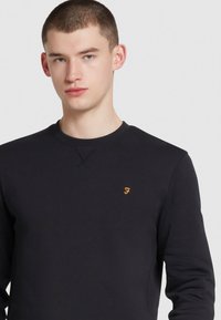 Granatowy sweter z bawełny z gładką strukturą, okrągłym dekoltem i małym, żółtym haftowanym logo po lewej stronie klatki piersiowej.