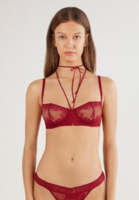 Czerwony biustonosz z koronką i siateczkowymi wstawkami, z regulowanymi ramiączkami i wiązanym detalem choker. Gładkie tekstury z kwiatowymi wzorami.