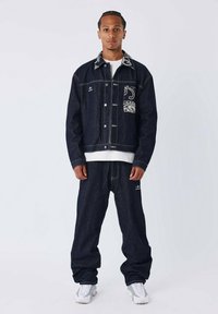 Donkerblauwe spijkerjas en -broek met witte stiksels, voorzien van een gemusterde zak, een relaxte pasvorm en witte sneakers.