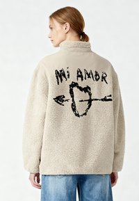Pullover in pile beige con collo alto; presenta una stampa grafica nera con la scritta "Mi Amor" e una freccia sulla schiena.