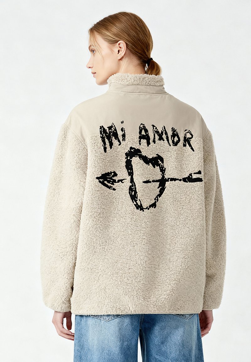 Pullover in pile beige con collo alto; presenta una stampa grafica nera con la scritta "Mi Amor" e una freccia sulla schiena.
