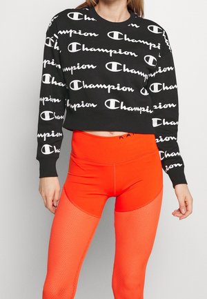 Kvinde iført sort cropped sweatshirt med hvide "Champion"-logoer og klare orange leggings i taljen med netpaneler.