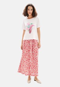 T-shirt blanc avec un imprimé d'oiseau coloré, associé à une longue jupe florale rouge. Texture de tissu lisse, design simple et chaussures de couleur neutre.