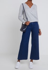 Top gris de manga larga con escote en V, vaqueros anchos azul marino, zapatillas blancas y bolso oscuro. La persona está de pie contra un fondo neutro.