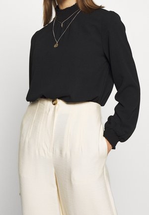 Zwarte blouse met een hoge, geplooide hals en pofmouwen, gecombineerd met crème wijde pijpenbroek met een voorknop en verticale strepen.