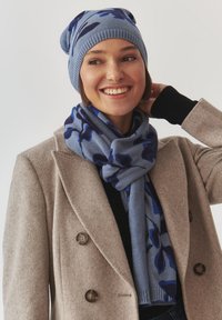 Ensemble bonnet et écharpe en tricot bleu avec un motif floral en bleu foncé. Texture douce, matériau chaud, associé à un manteau beige.