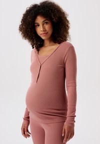 Gravid kvinna med lockigt hår som bär en figurn ära långärmad topp i grårosa och matchande byxor, stående mot en vit bakgrund.