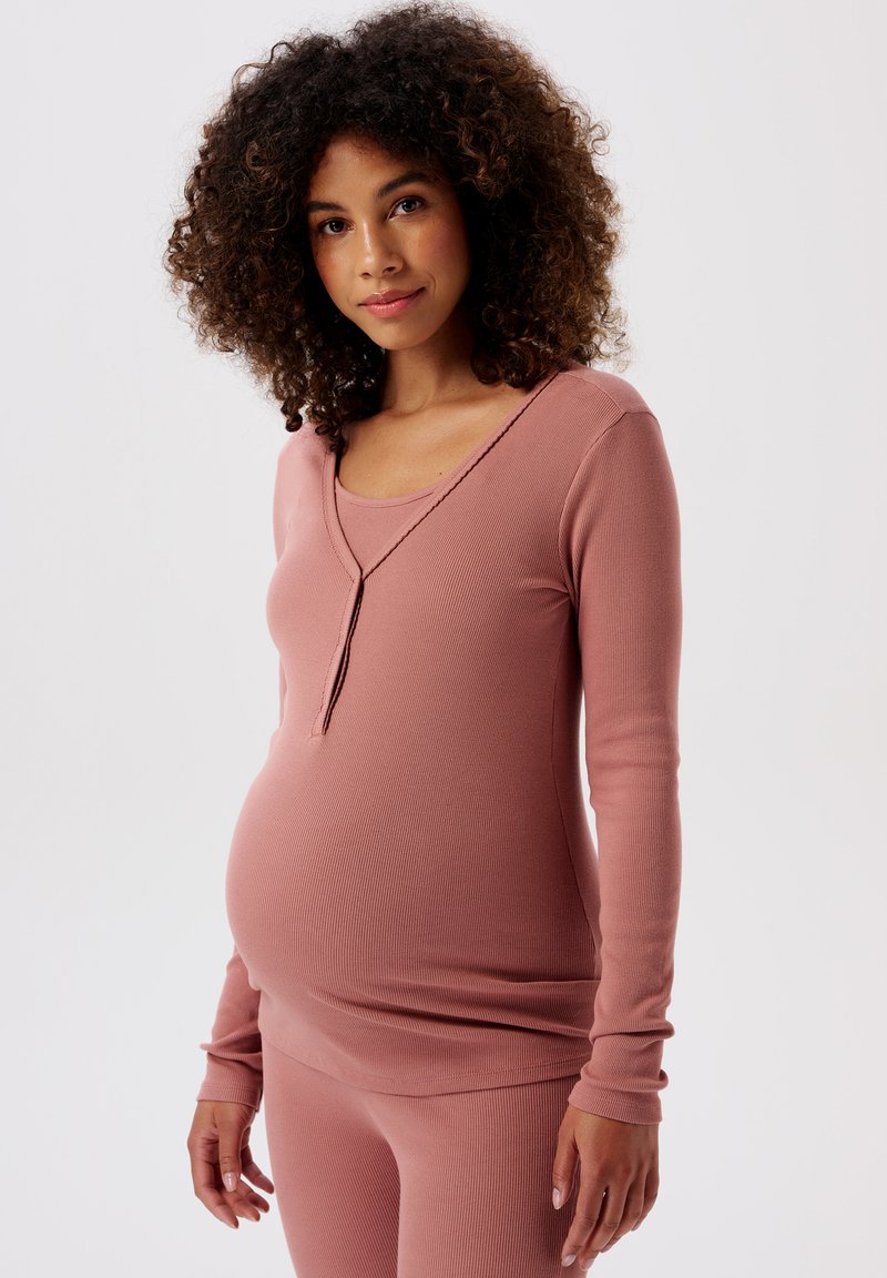 Gravid kvinna med lockigt hår som bär en figurnära långärmad topp i grårosa och matchande byxor, stående mot en vit bakgrund.