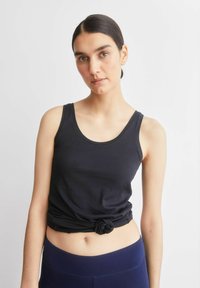 Zwart tanktopje met een ronde halslijn en een geknoopt zoom. De stof lijkt glad en aansluitend, gecombineerd met donkerblauwe leggings.