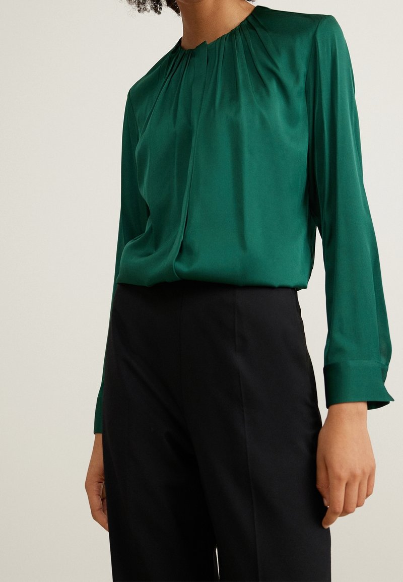 Femme portant un chemisier vert émeraude à manches longues avec des plis au niveau de l'encolure et un pantalon noir taille haute sur un fond uni.