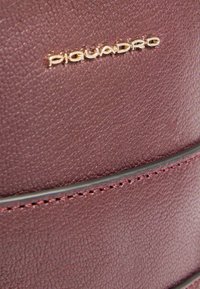 Borsa in pelle bordeaux con superficie texturizzata, marchio "Piquadro" in oro e cuciture scure a contrasto lungo una linea di accento liscia.