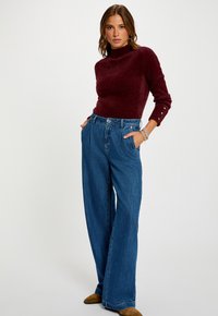 Donna in piedi con le mani in tasca, che indossa un maglione lungo bordeaux, jeans blu a gamba larga, scarpe marroni e braccialetti d'argento.