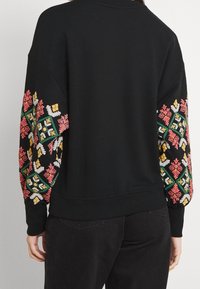 Czarny longsleeve z kolorowym haftem kwiatowym na rękawach, noszony przez osobę zwróconą plecami, w połączeniu z czarnymi spodniami.