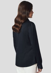 Granatowa bluzka z długim rękawem wykonana z gładkiego materiału, posiadająca dopasowany krój, detale w postaci plis na plecach oraz zapięcie na guziki przy mankietach.