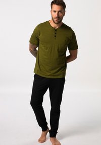 Camisa henley de manga corta en verde oliva con tres botones negros y un bolsillo en el pecho, combinada con pantalones negros ajustados. Corte casual, textura suave.