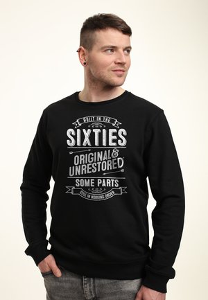 Mężczyzna w czarnej bluzie z białym napisem: "Zbudowany w latach sześćdziesiątych, oryginalny i nieodnowiony, niektóre części nadal działają."