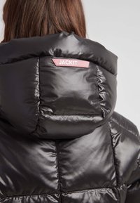 Chaqueta acolchada negra con un acabado brillante. Presenta un cuello alto, un diseño geométrico acolchado y un parche de logo rosa en la parte trasera.