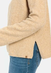 Beige strikket sweater med en blød tekstur, der har slidser i siderne og et lille mærketag. Kombineret med blå denim jeans.