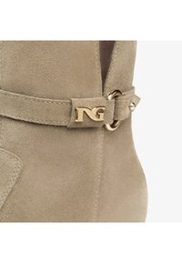 Bottine en daim beige présentant un design épuré, des accessoires en métal doré, un détail de boucle circulaire et une ouverture latérale.