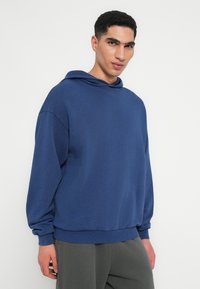 Kék pamut kapucnis pulóver, zsinórral, bordázott mandzsettákkal és alsó résszel, laza fitccel, a mellkason egy diszkrét logóval. Puha textúra, minimális design.