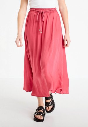 Donna che indossa una gonna midi corallo fluente con vita regolabile a coulisse e sandali neri con fibbia borchiata mentre cammina in avanti.