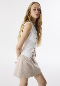 Top blanco sin mangas con textura acanalada y cierres de botón plateados, combinado con una falda mini texturizada beige con bolsillos.