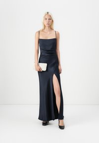 Ναυτικό σατέν βραδινό φόρεμα με λεπτές τιράντες, εφαρμοστό μπούστο και ψηλή σχισμή. Το μοντέλο κρατάει μια κρουστική λευκή τσάντα, συνδυασμένη με μαύρα παπούτσια.