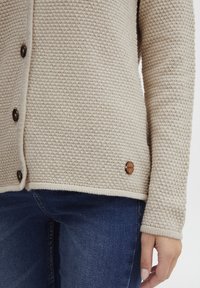 Beżowy dzianinowy kardigan z fakturowanym wzorem, z ciemnymi guzikami i drewnianym logo na dolnym brzegu. Noszony na niebieskich dżinsach.