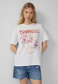 T-shirt blanc en coton à manches courtes, présentant un graphisme de fleurs tropicales et de palmiers. Inclut du texte dans des tons rouges et roses.