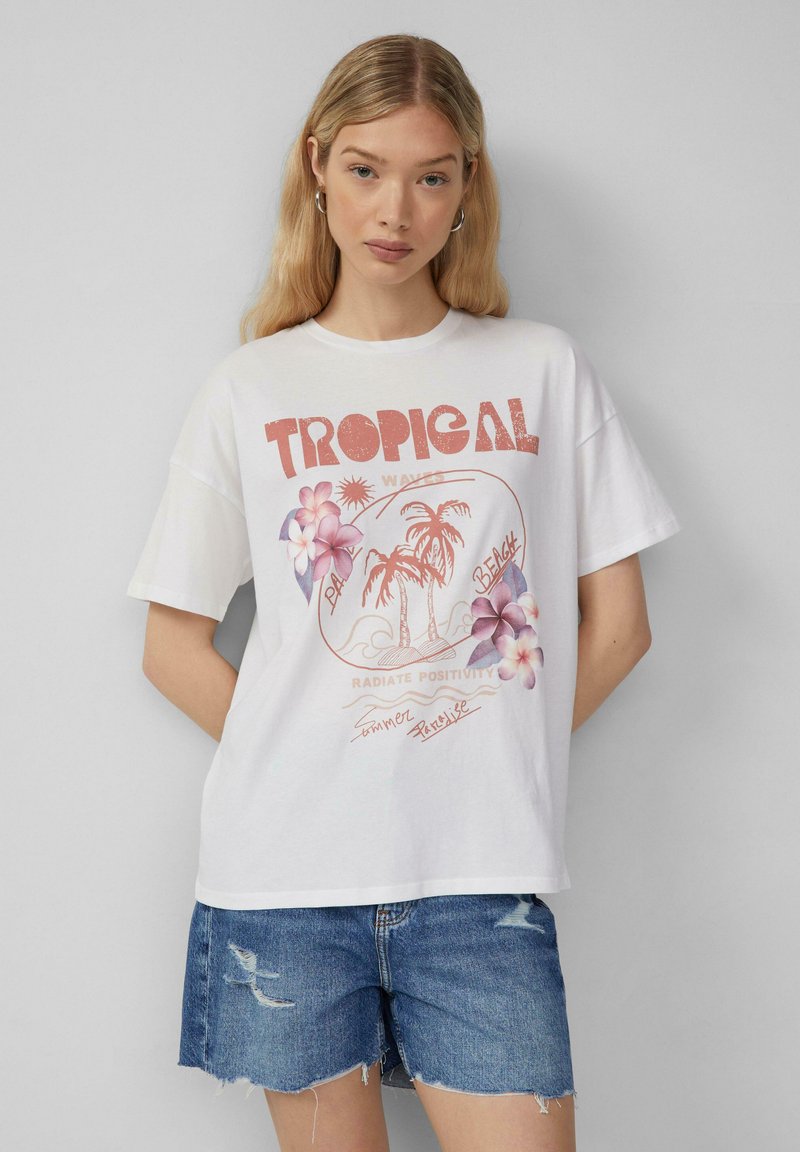 T-shirt blanc en coton à manches courtes, présentant un graphisme de fleurs tropicales et de palmiers. Inclut du texte dans des tons rouges et roses.