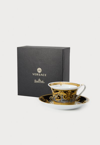Φλιτζάνι τσαγιού από πορσελάνη Versace με περίτεχνο μαύρο και χρυσό σχέδιο, συνοδευόμενο από αντίστοιχο πιάτο, παρουσιάζεται σε μαύρο κουτί με λογότυπο.