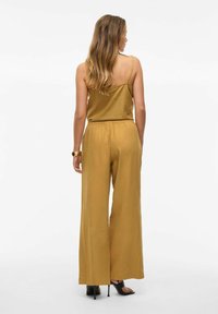 Mono dorado amarillento con un corte holgado, tirantes tipo espagueti, cintura elástica y pantalones de pierna ancha. Combinado con sandalias negras de tacón.