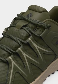 Κοντινό πλάνο αθλητικού παπουτσιού σε ελαιί πράσινο με υφασμάτινη υφή, ζιγκ-ζαγκ συνθετικές επενδύσεις, κορδόνια με σχέδιο και μπεζ σόλα.